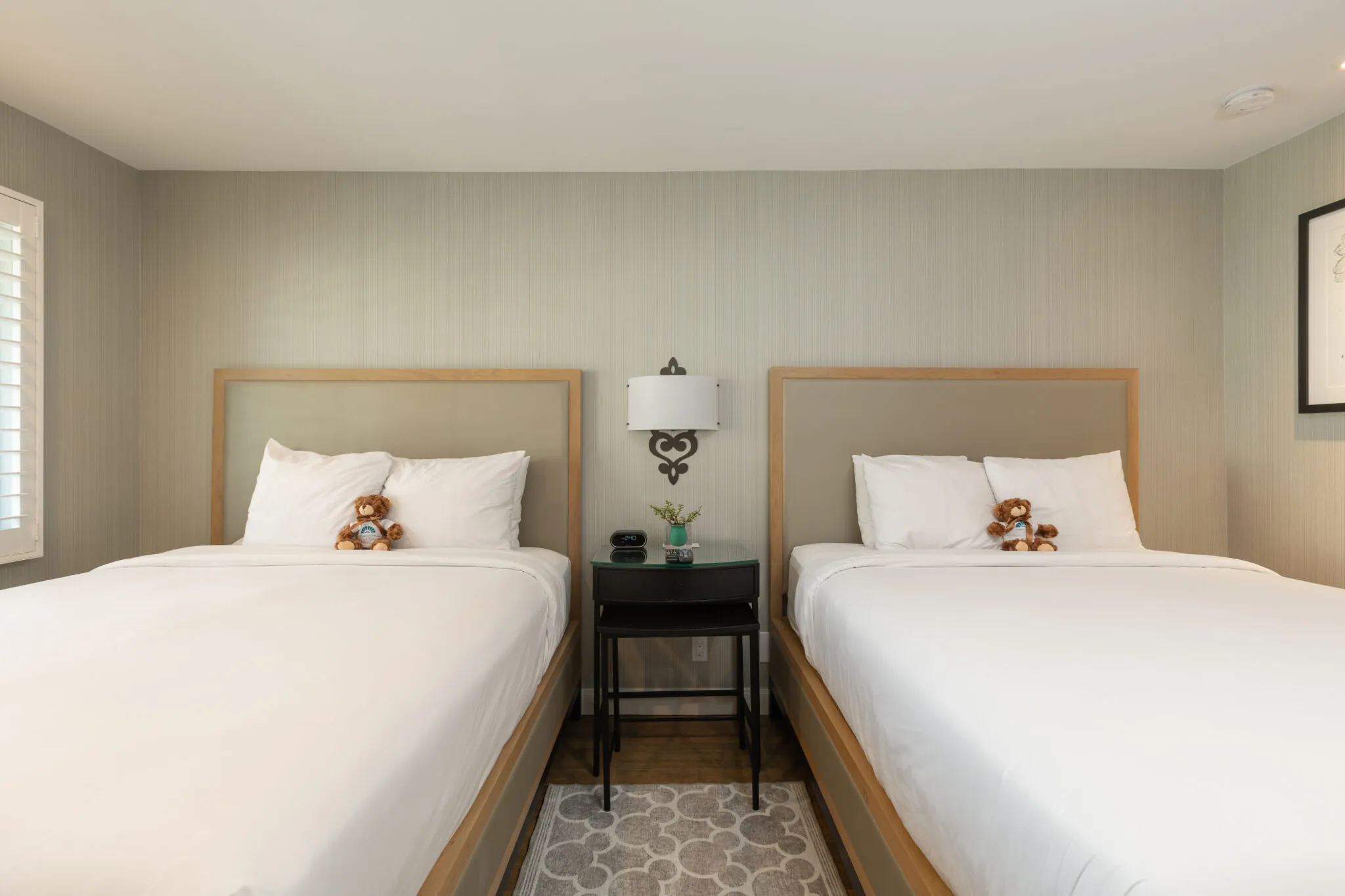 A hotel room with two beds, a nightstand, and teddy bears on the pillows.